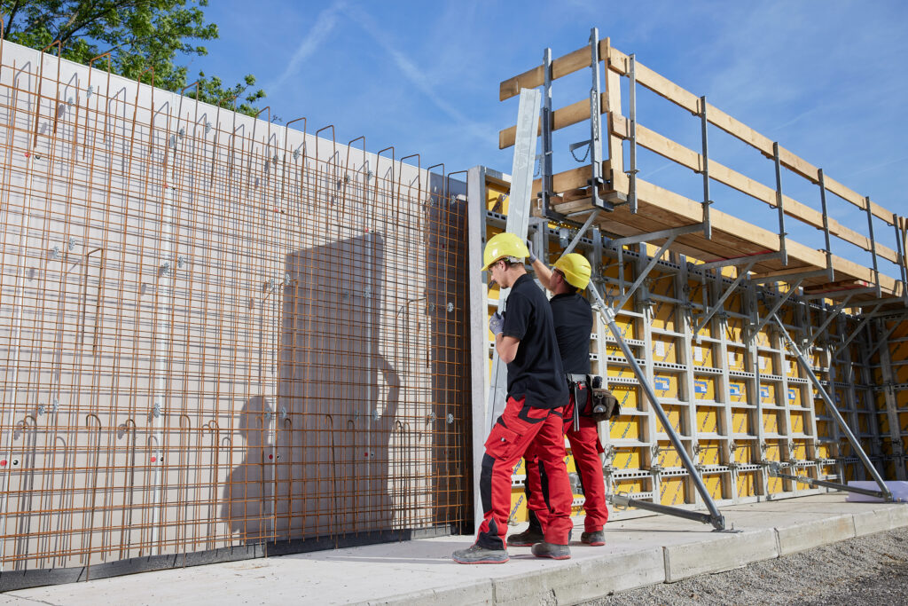 Zwei Personen mit gelben Schutzhelmen richten eine Schalungsstütze an einer bewehrten Betonwand auf einer Baustelle aus.