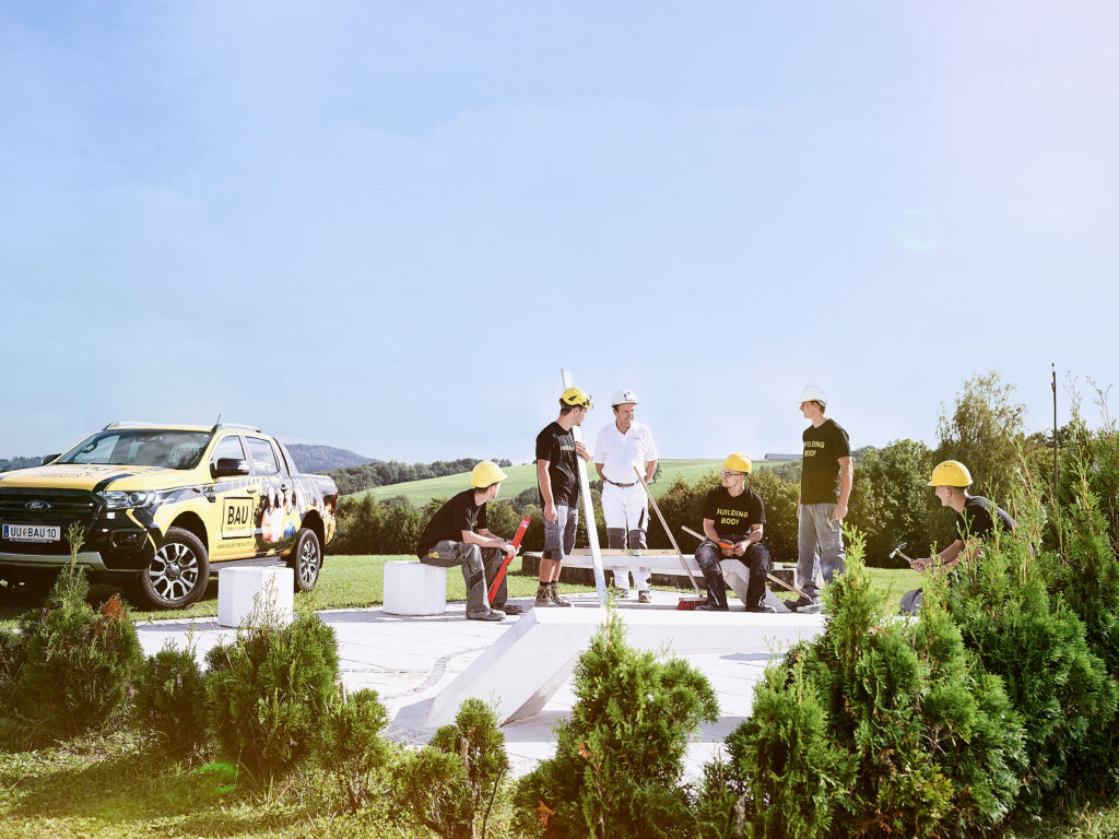 Gruppe von Personen mit Schutzhelmen arbeitet und bespricht Aufgaben auf einer Baustelle im Freien; links steht ein gelbes BAU-Akademie-Fahrzeug.