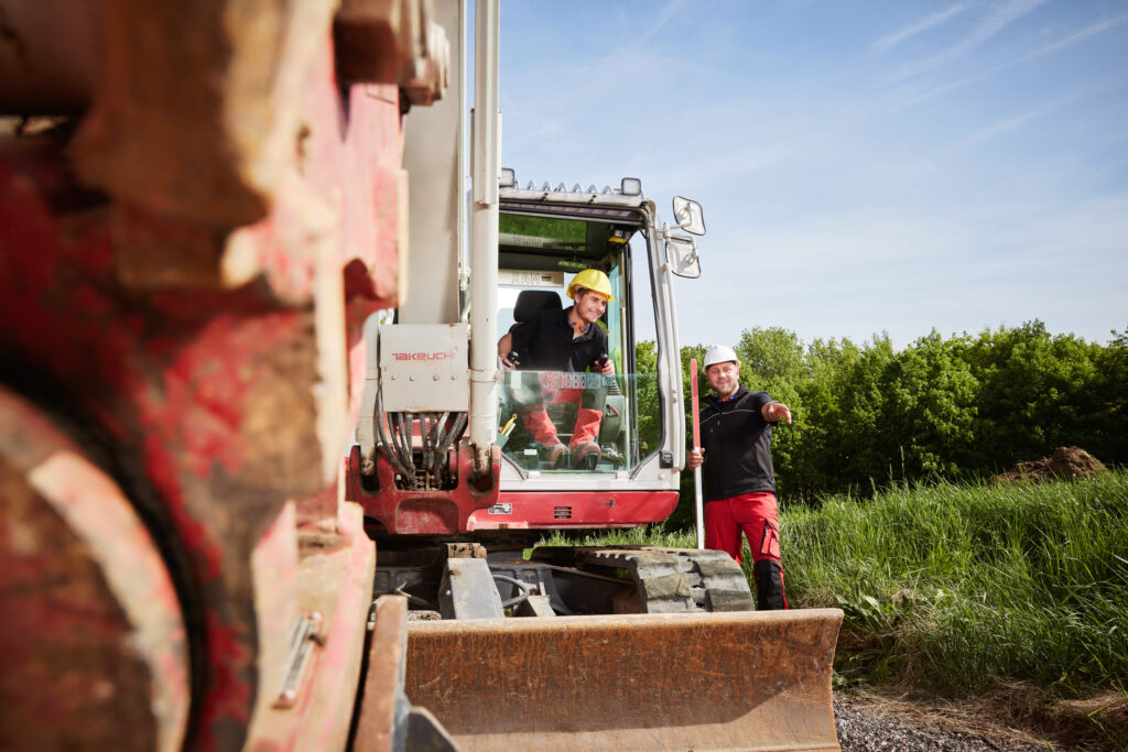 Person im Bagger lächelt aus der Kabine, während eine zweite Person mit Schutzhelm danebensteht und in eine Richtung zeigt