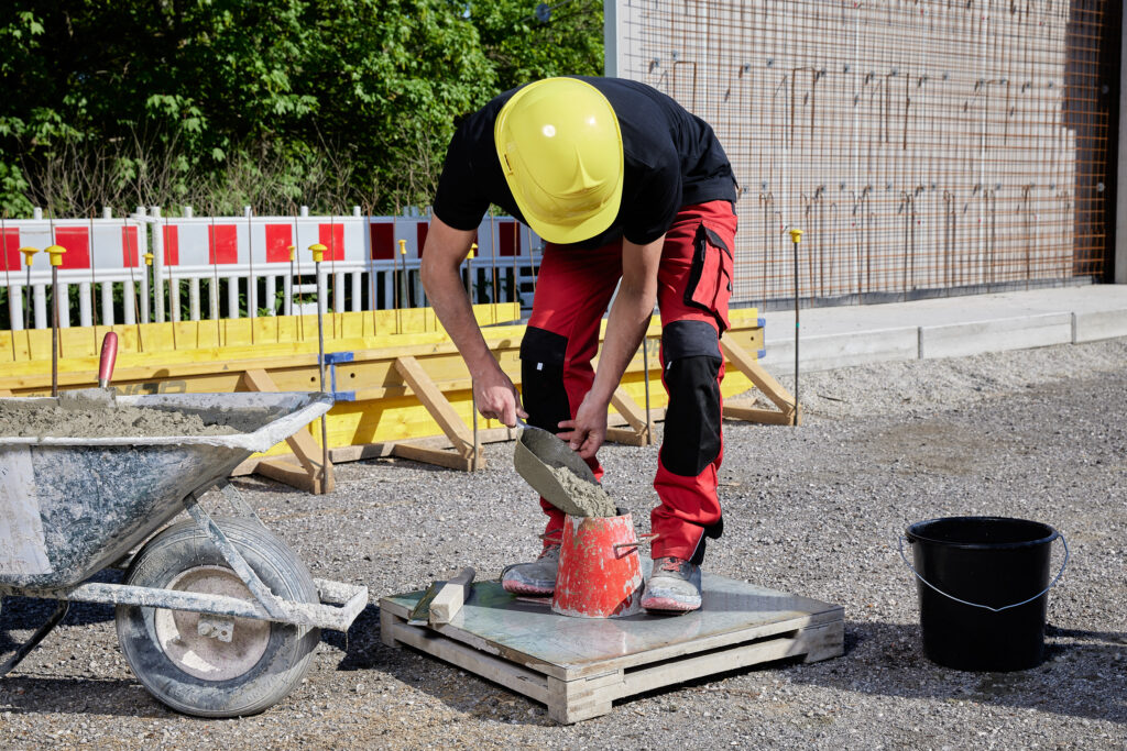 Person mit gelbem Schutzhelm füllt frischen Beton mit einer Kelle in einen roten Prüfkegel; daneben Schubkarre und Wassereimer