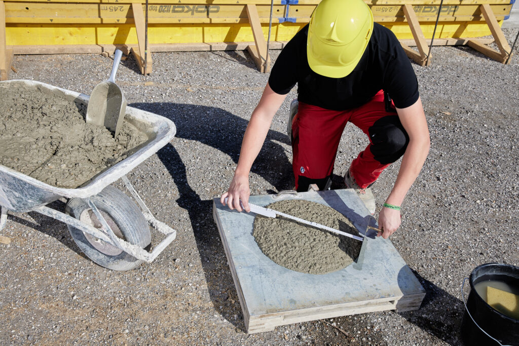 Person mit Schutzhelm misst die Ausbreitung von frischem Beton auf einer Prüffläche; daneben Schubkarre mit Beton und Wassereimer.