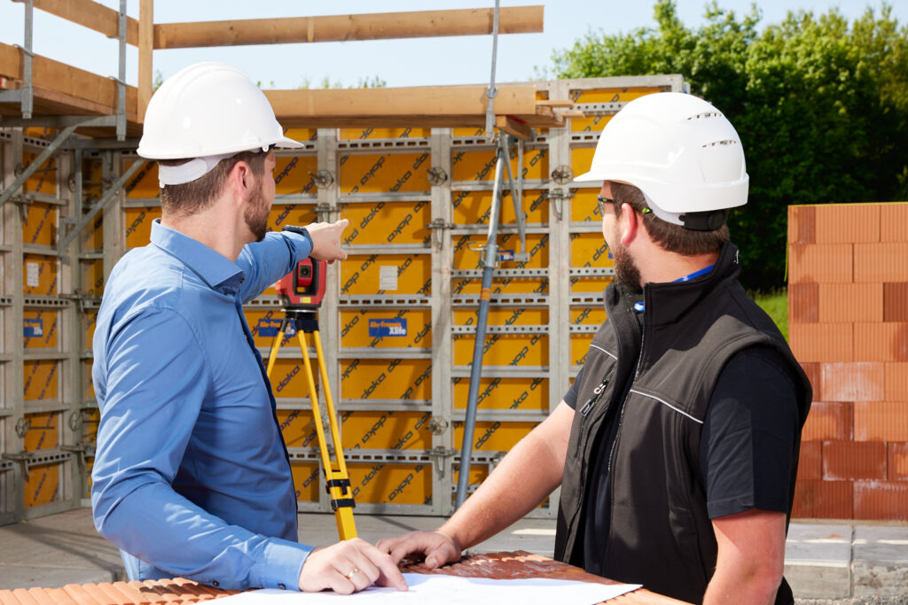 „Zwei Personen mit weißen Schutzhelmen besprechen Baupläne; eine zeigt auf die Schalungskonstruktion im Hintergrund, neben einem Vermessungsstativ.