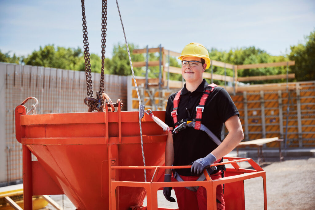 Person mit Schutzhelm und Sicherheitsgurt steht auf einer Arbeitsbühne neben einem roten Betonbehälter auf einer Baustelle.