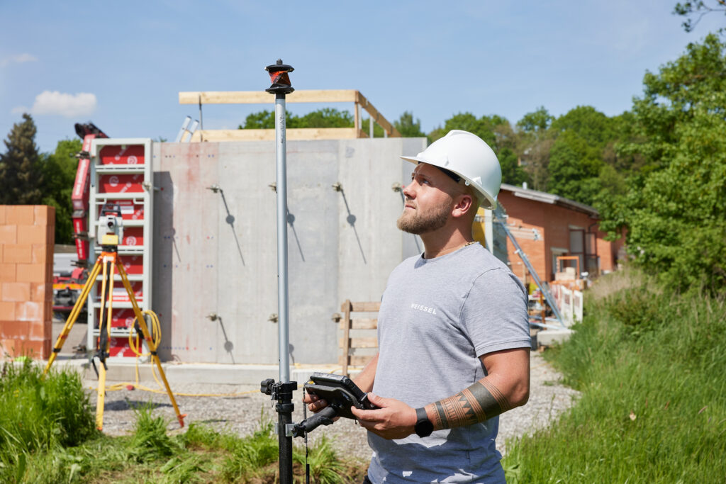Person mit weißem Schutzhelm führt Vermessungsarbeiten durch und hält ein Messgerät; im Hintergrund Baustelle mit Betonwand und Vermessungsstativ