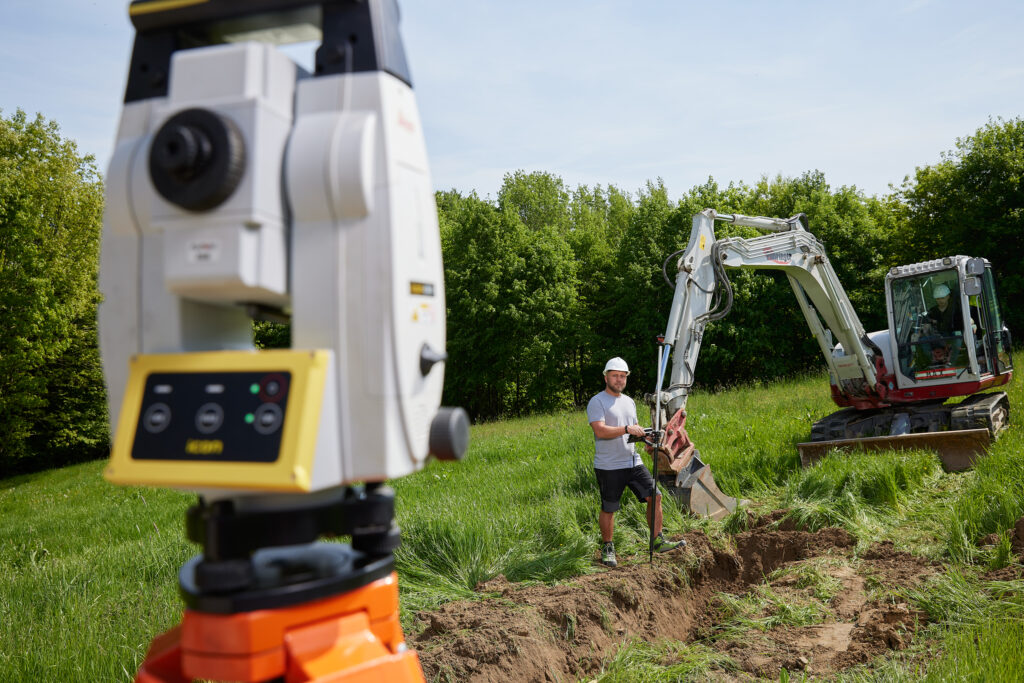 Vermessungsgerät im Vordergrund; im Hintergrund steht eine Person mit Schutzhelm in einem ausgehobenen Graben neben einem Bagger.