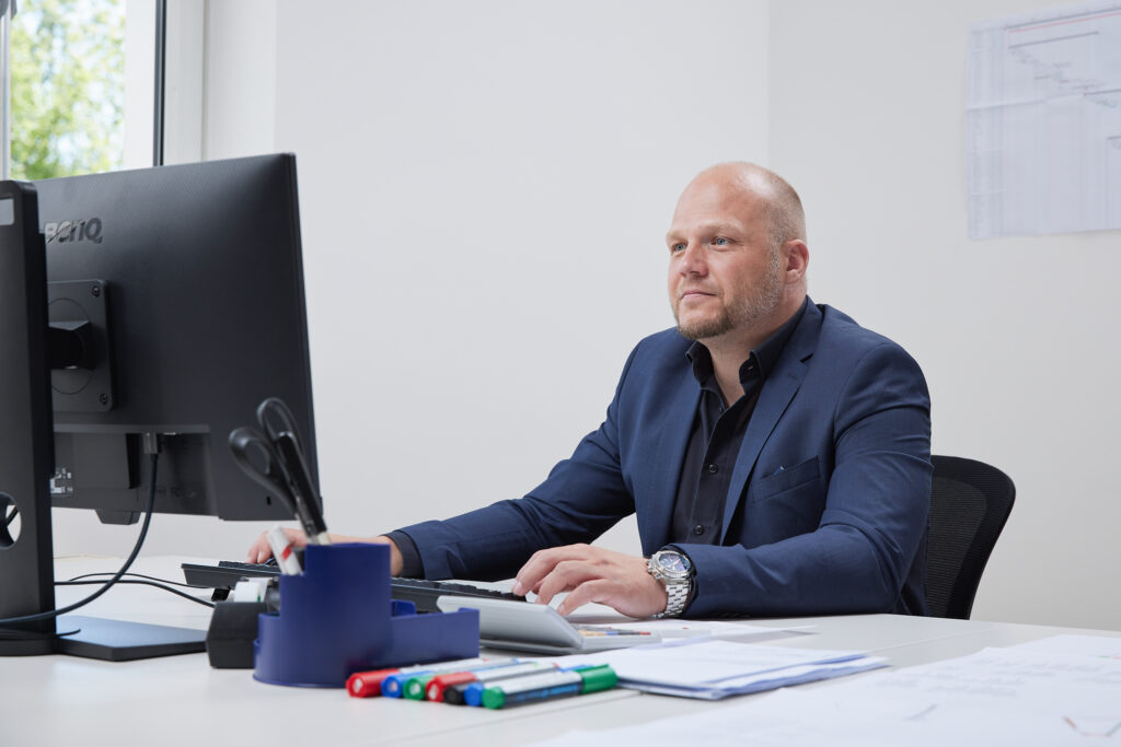 Person im Büro sitzt konzentriert vor einem Computerarbeitsplatz; Schreibtisch mit Unterlagen und Stiften im Vordergrund.