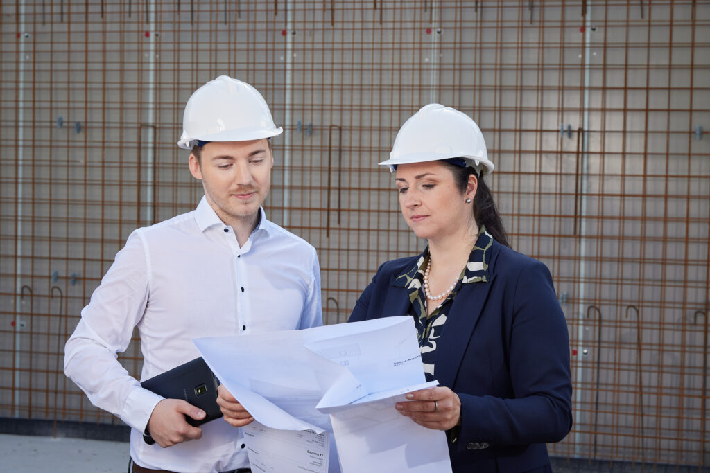 Zwei Personen mit Schutzhelmen betrachten gemeinsam Baupläne auf einer Baustelle.