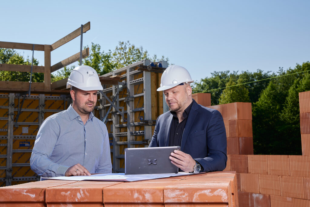 Zwei Personen mit weißen Schutzhelmen besprechen Baupläne und schauen gemeinsam auf ein Tablet auf einer Baustelle.