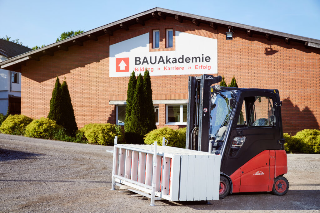 Gabelstapler transportiert ein Metallgestell vor einem Gebäude mit BAUAkademie-Beschilderung.
