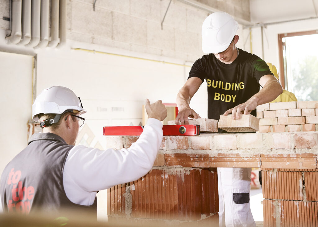 Zwei Personen mit Schutzhelmen arbeiten an einer Mauer; eine Person richtet eine Wasserwaage aus, die andere setzt Ziegelsteine