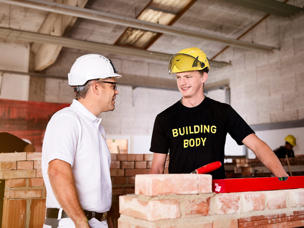 Zwei Personen mit Schutzhelmen unterhalten sich an einer gemauerten Wand; eine Person lächelt und hält eine Wasserwaage.