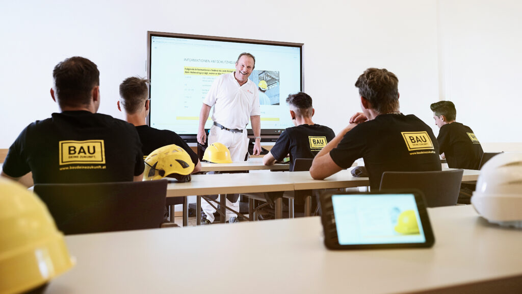 Lehrperson steht vor einer Präsentation und unterrichtet eine Gruppe junger Menschen in einem Klassenraum; gelbe Schutzhelme liegen auf den Tischen